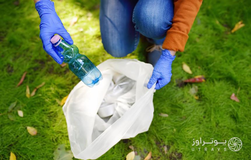 یک جفت دستِ دستکش‌پوشیده زنانه درحال انداختن یک بطری آبِ خالی داخل کیسه زباله